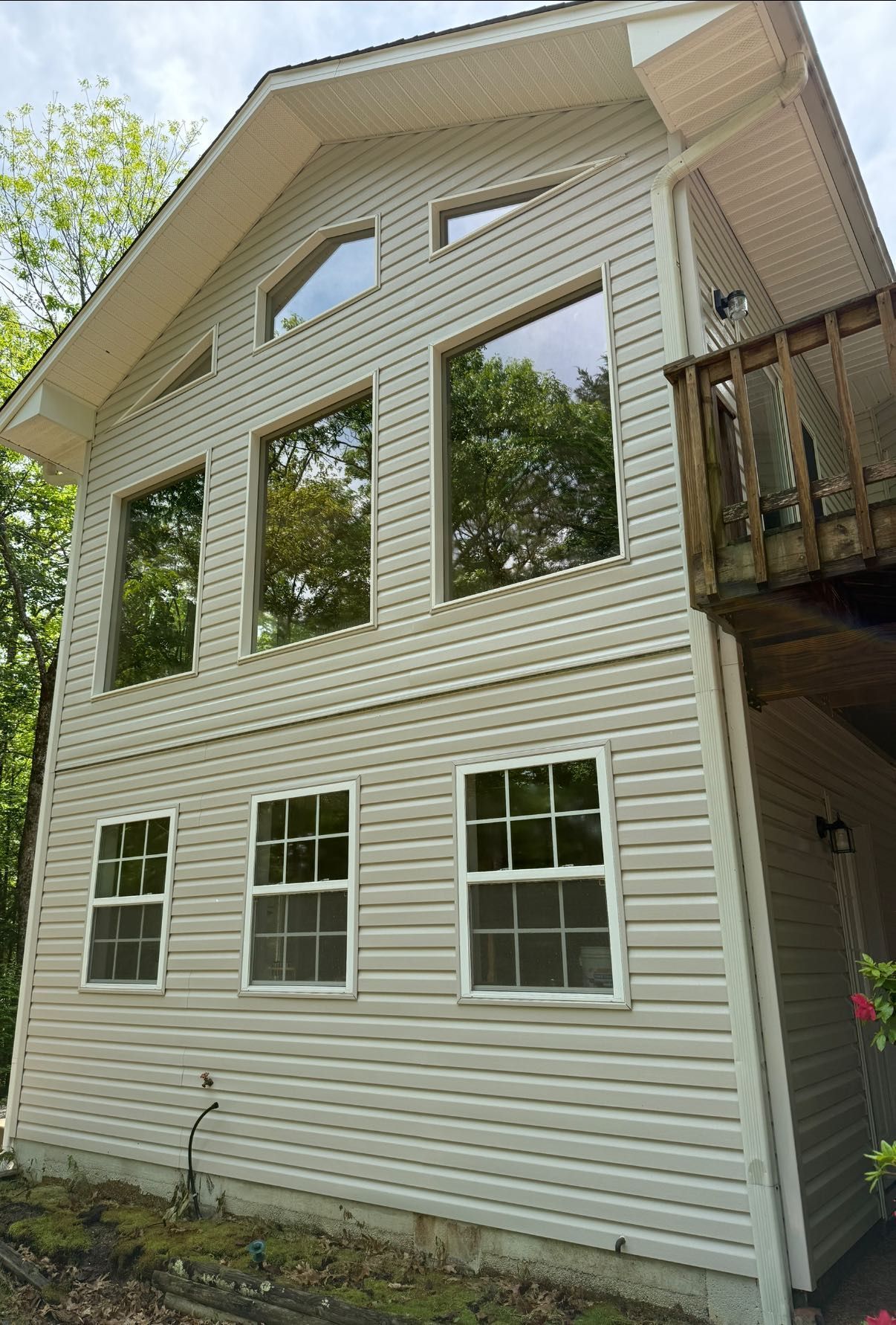 Front view of a house with beige siding, dark shingle roof, and two windows under trees and blue sky