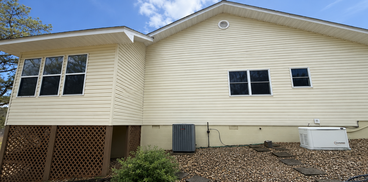 Side view of a beige house with white trim, windows, a wooden deck, and utility units on the ground.