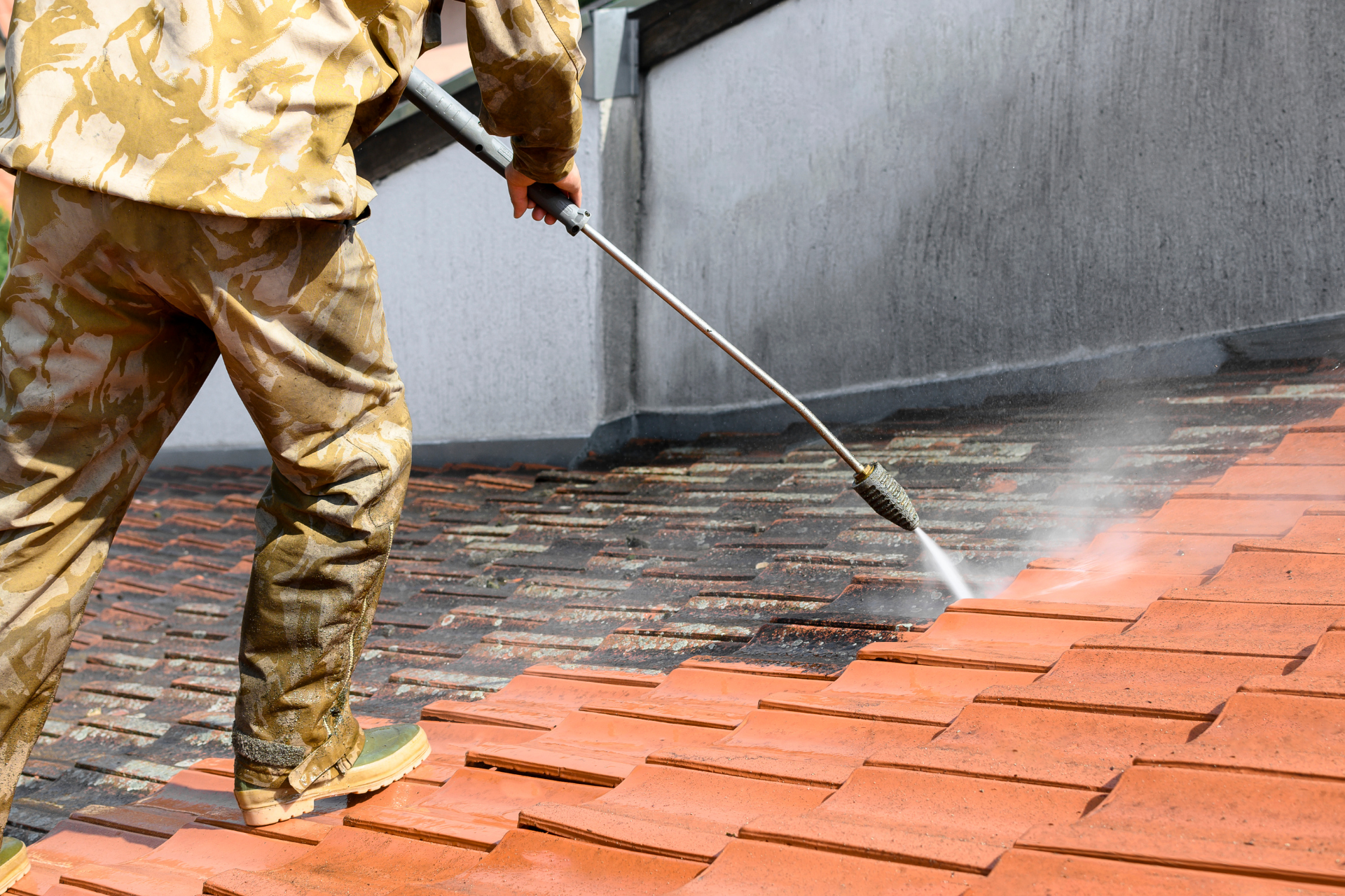 Worker pressure-washing red roof tiles with a hose, spraying water along a rooftop edge