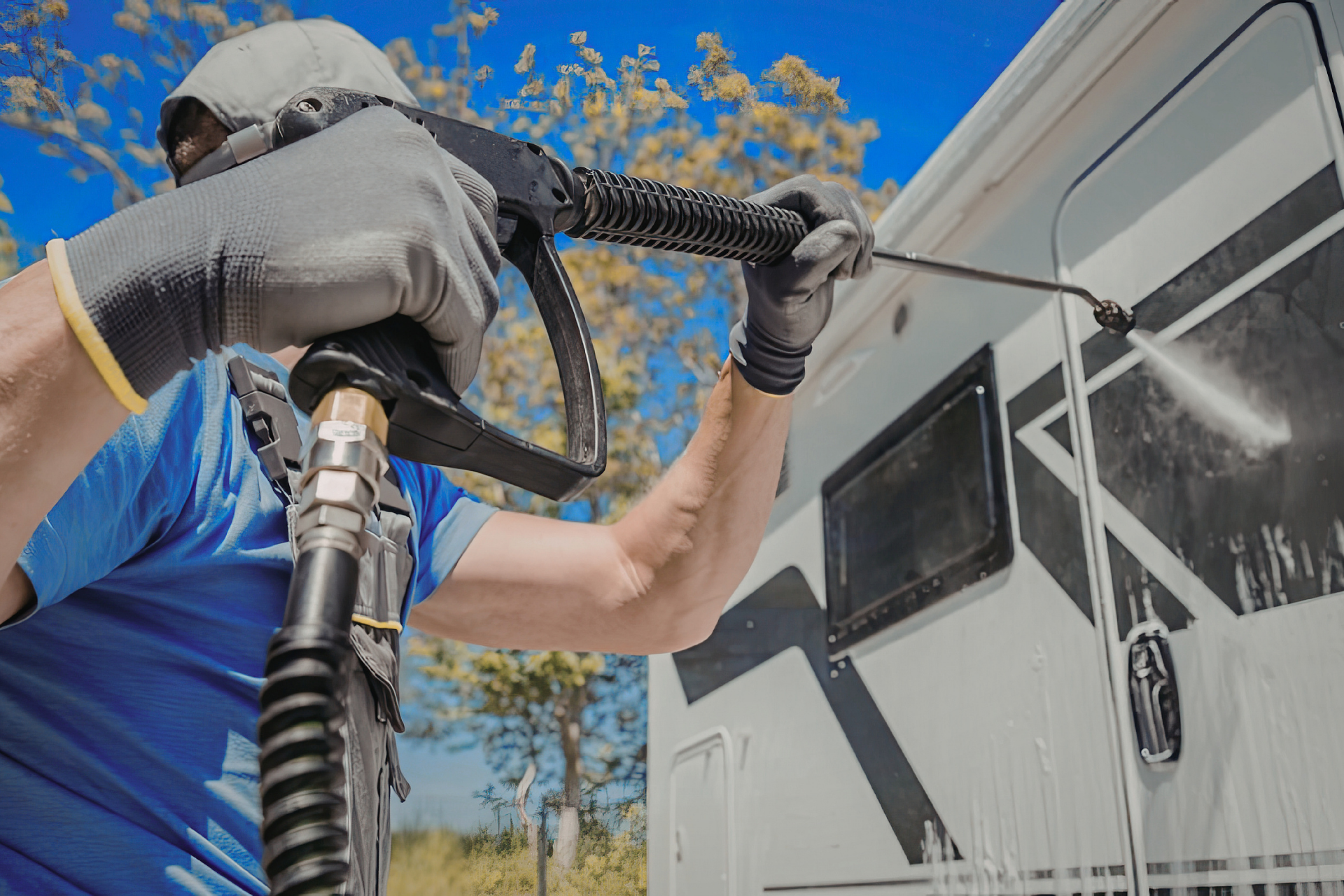 Worker pressure-washing a white RV outdoors with a black hose and spray nozzle