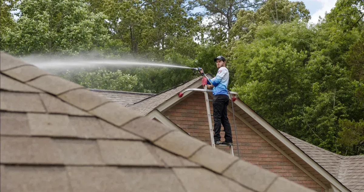 Worker pressure-washing a shingled roof on a house surrounded by trees