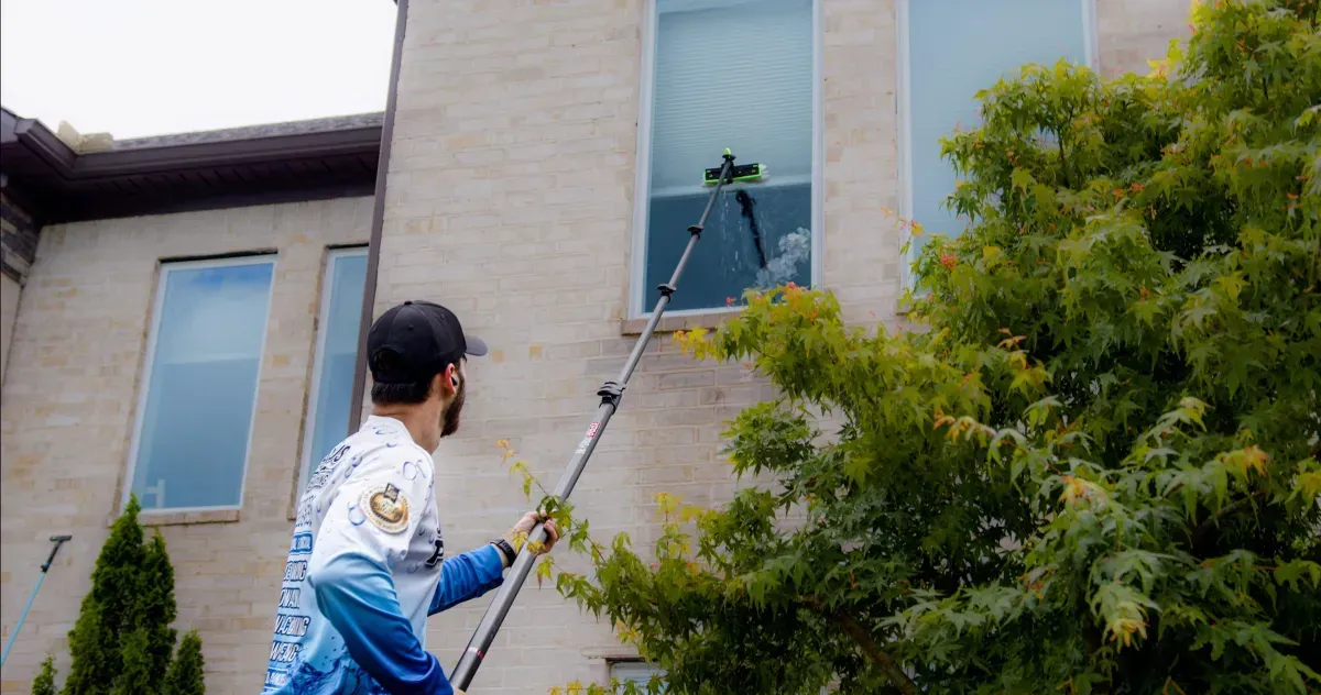 Worker using a pole washer cleans a window on the side of a brick building.