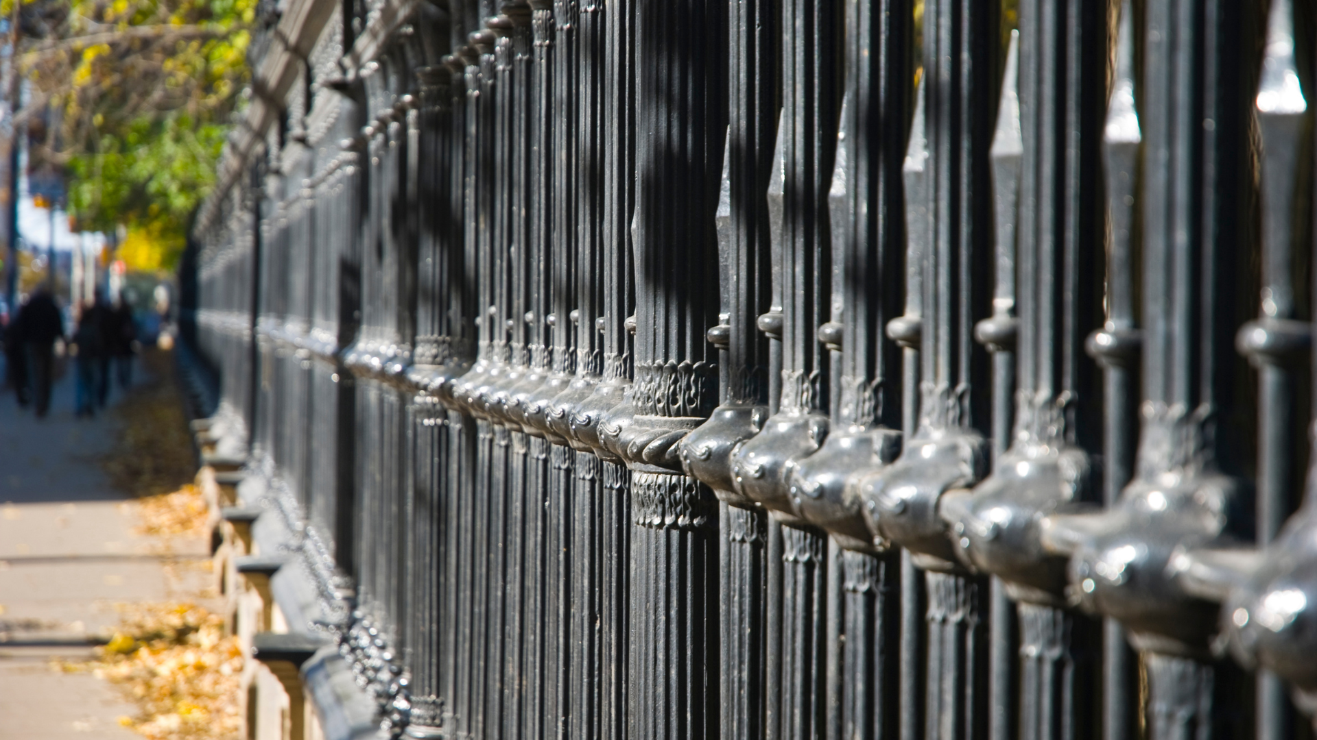 A close up of a wrought iron fence with people walking in the background.