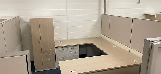 A cubicle with a desk , cabinets , drawers and a refrigerator.