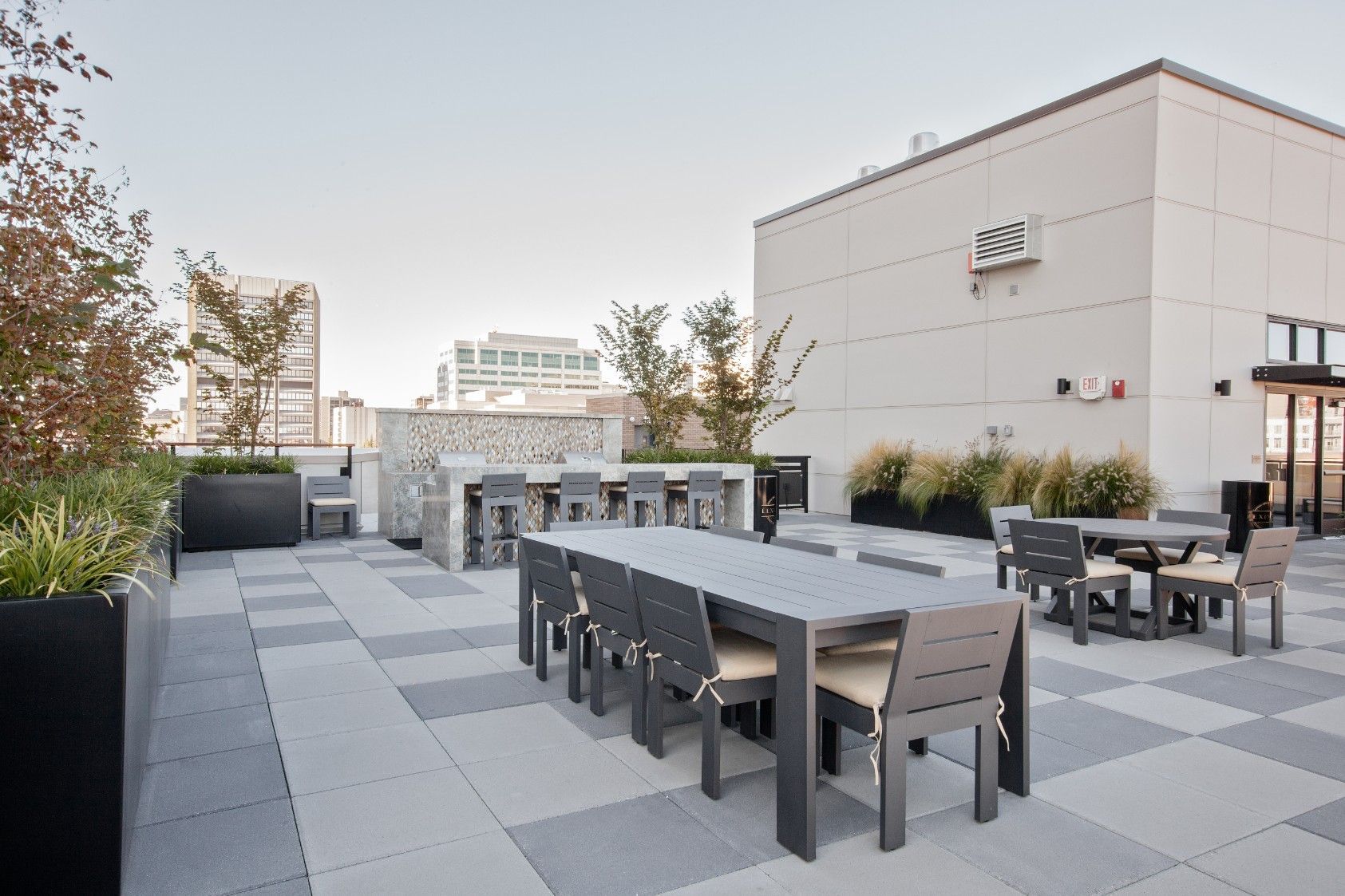 Rooftop patio with gray tables and chairs, potted plants, and city buildings in the background
