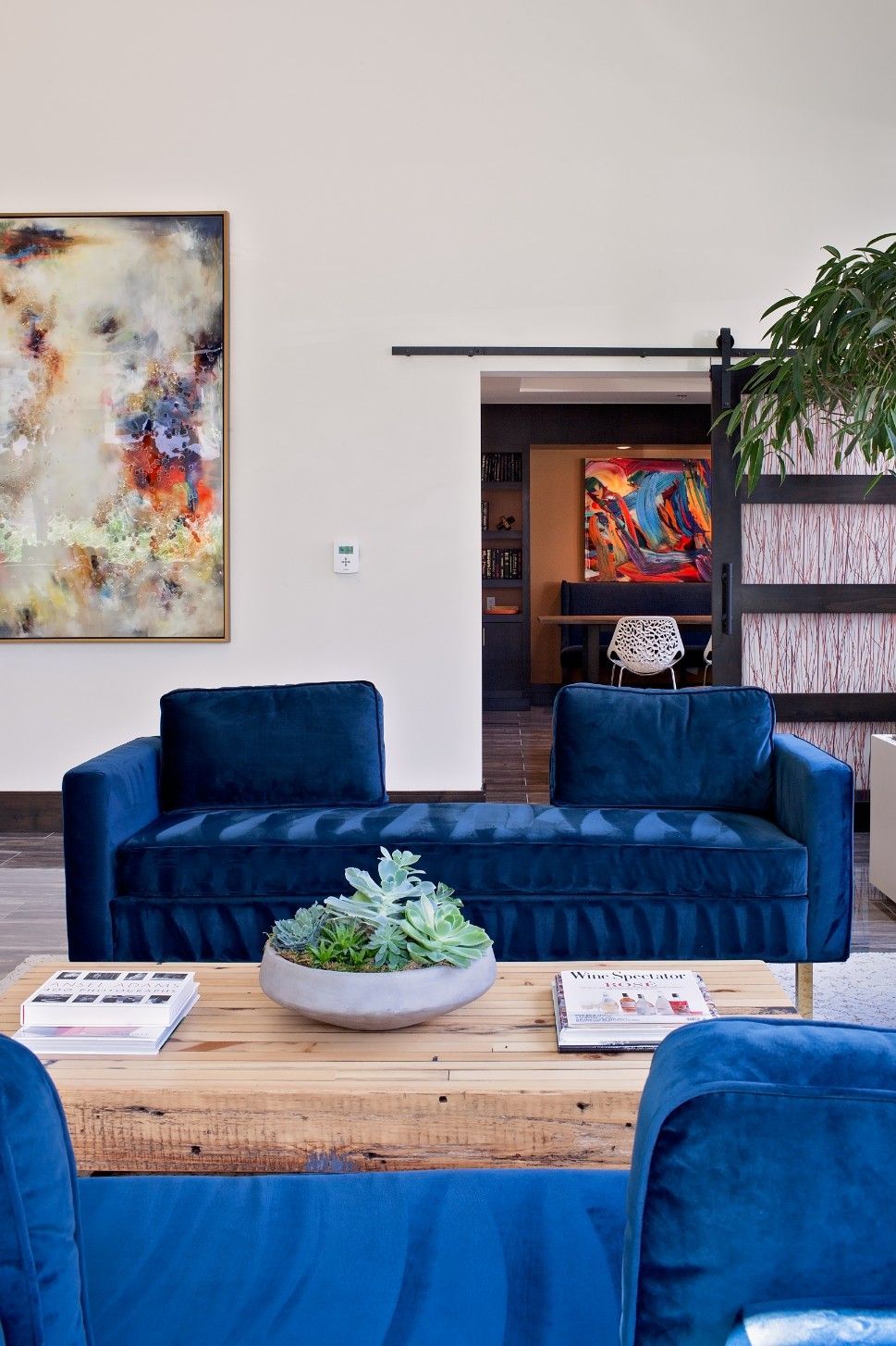 Modern living room with blue velvet sofas, wooden coffee table, abstract art, and a doorway to a colorful room