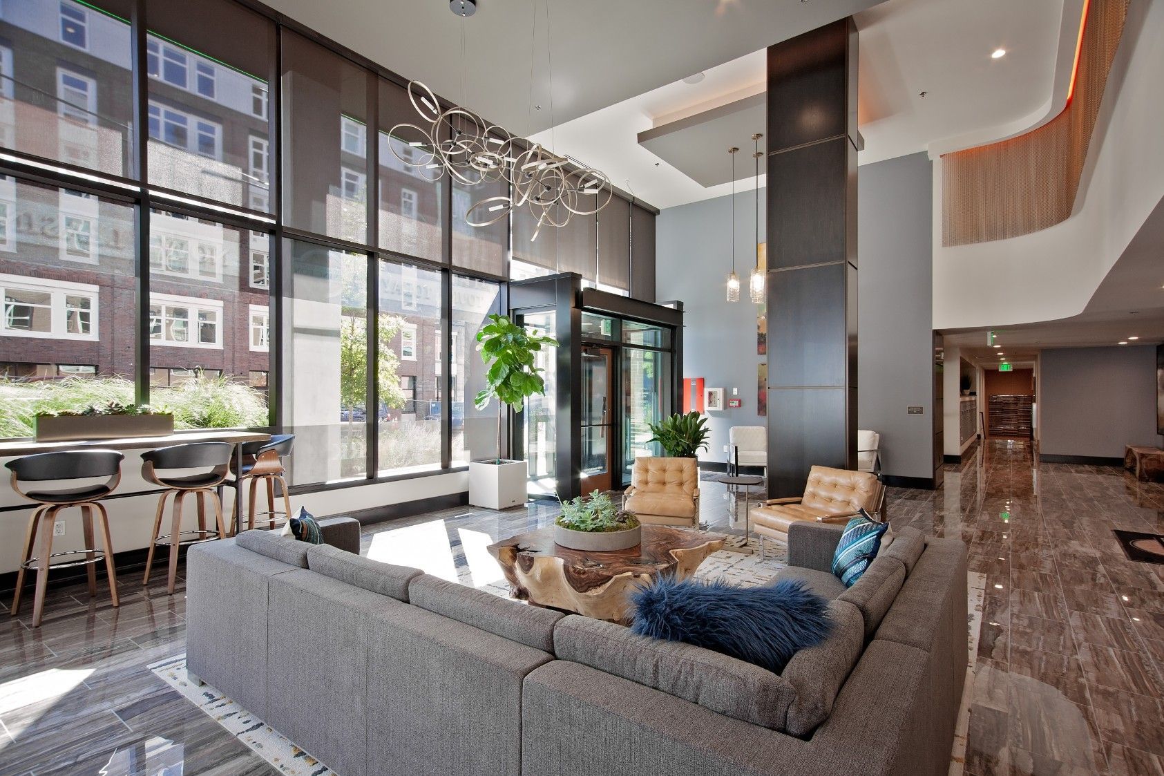 Modern apartment lobby with large windows, gray sectional sofa, dining tables, and tall columns