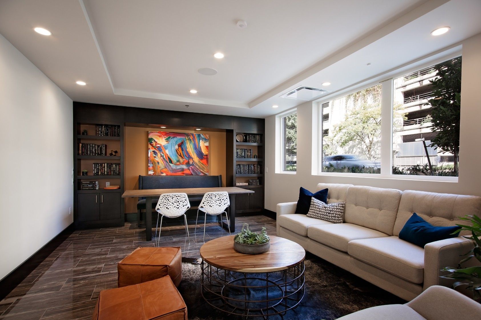 Modern living room with beige sofa, round coffee table, desk, and colorful wall art by large windows