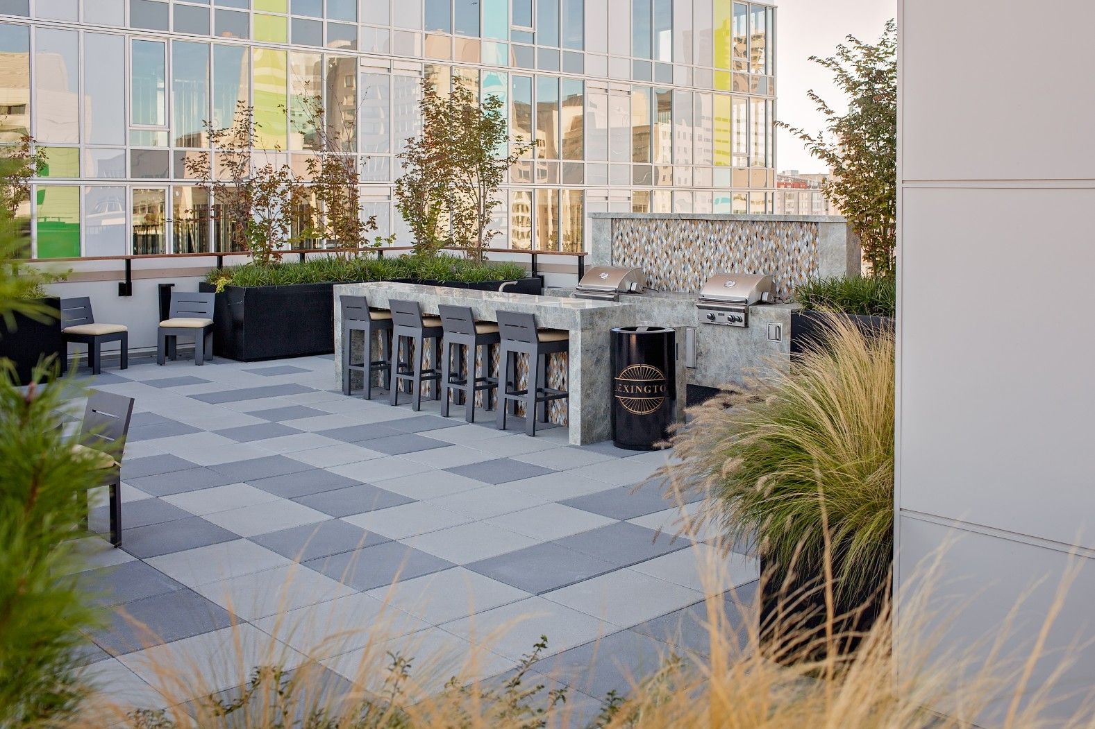 Outdoor patio with patterned pavers, bar stools, and tables beside a modern glass building.