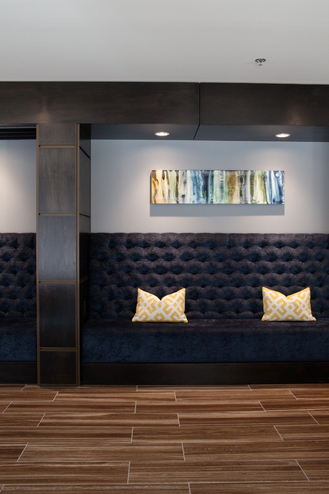 Modern lobby seating area with dark blue tufted bench, yellow pillows, and abstract wall art above wood flooring
