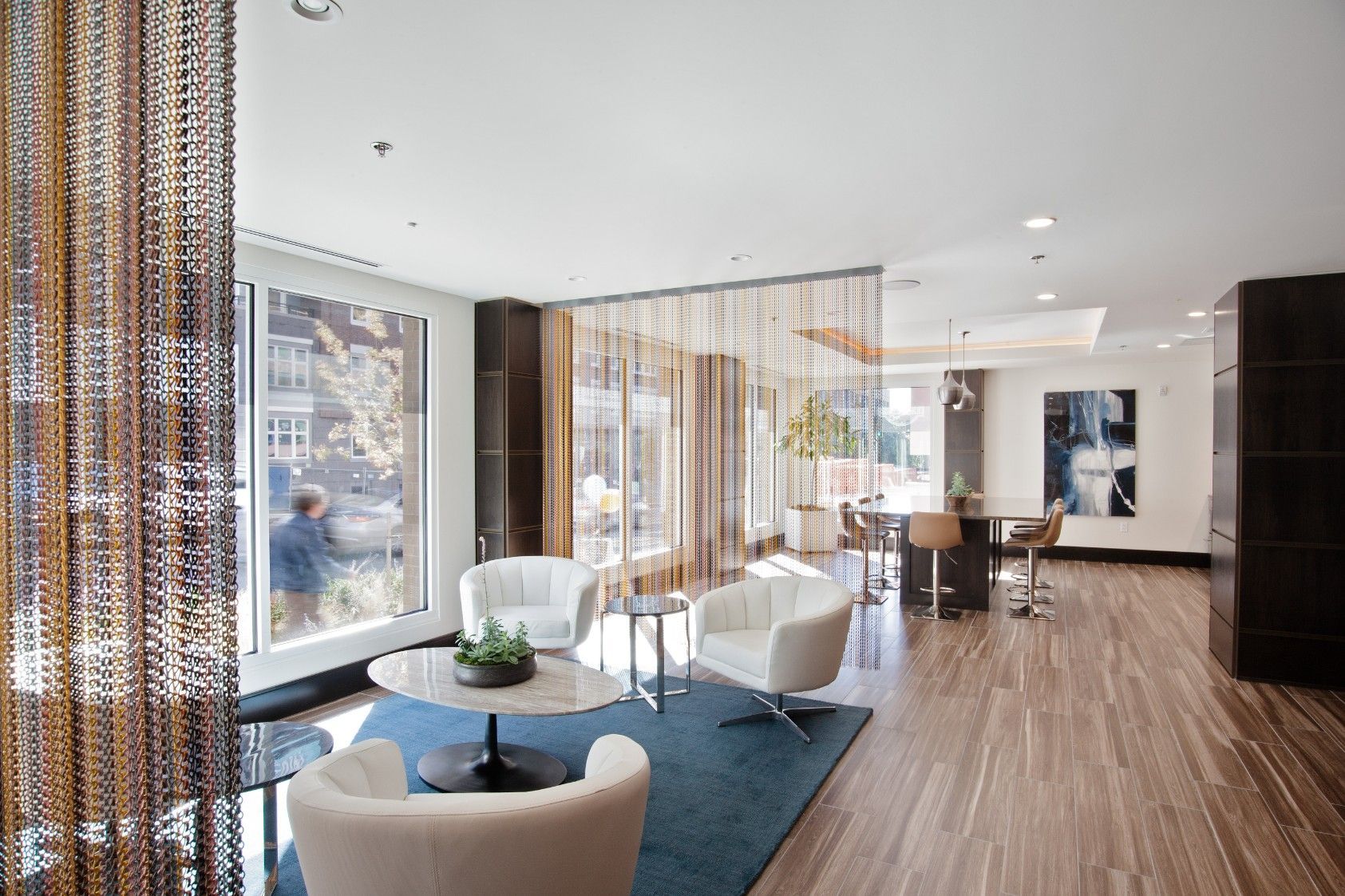 Modern living room with blue rug, white chairs, beaded curtains, and large windows overlooking city view