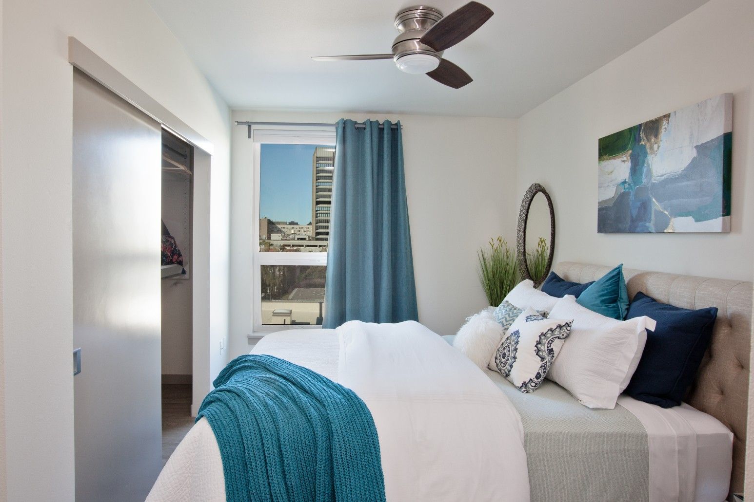 Bright bedroom with teal accents, white bedding, and a window view of city buildings.