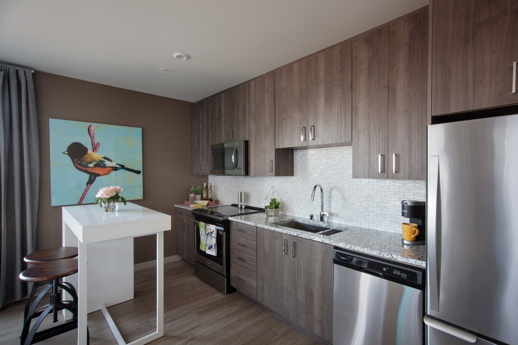 Modern kitchen with wood cabinets, stainless steel appliances, and a small white table by a bird painting