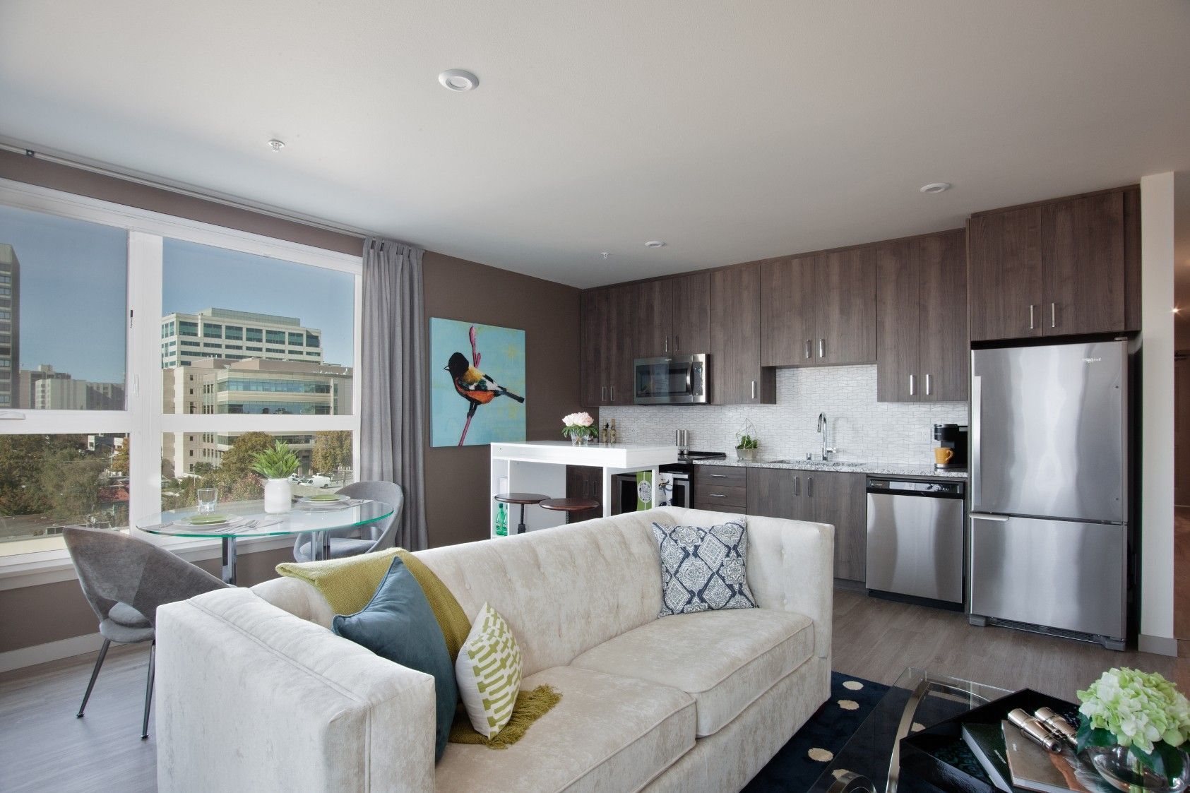Modern living room and kitchen with gray cabinets, white sofa, city-view windows, and a stainless steel fridge