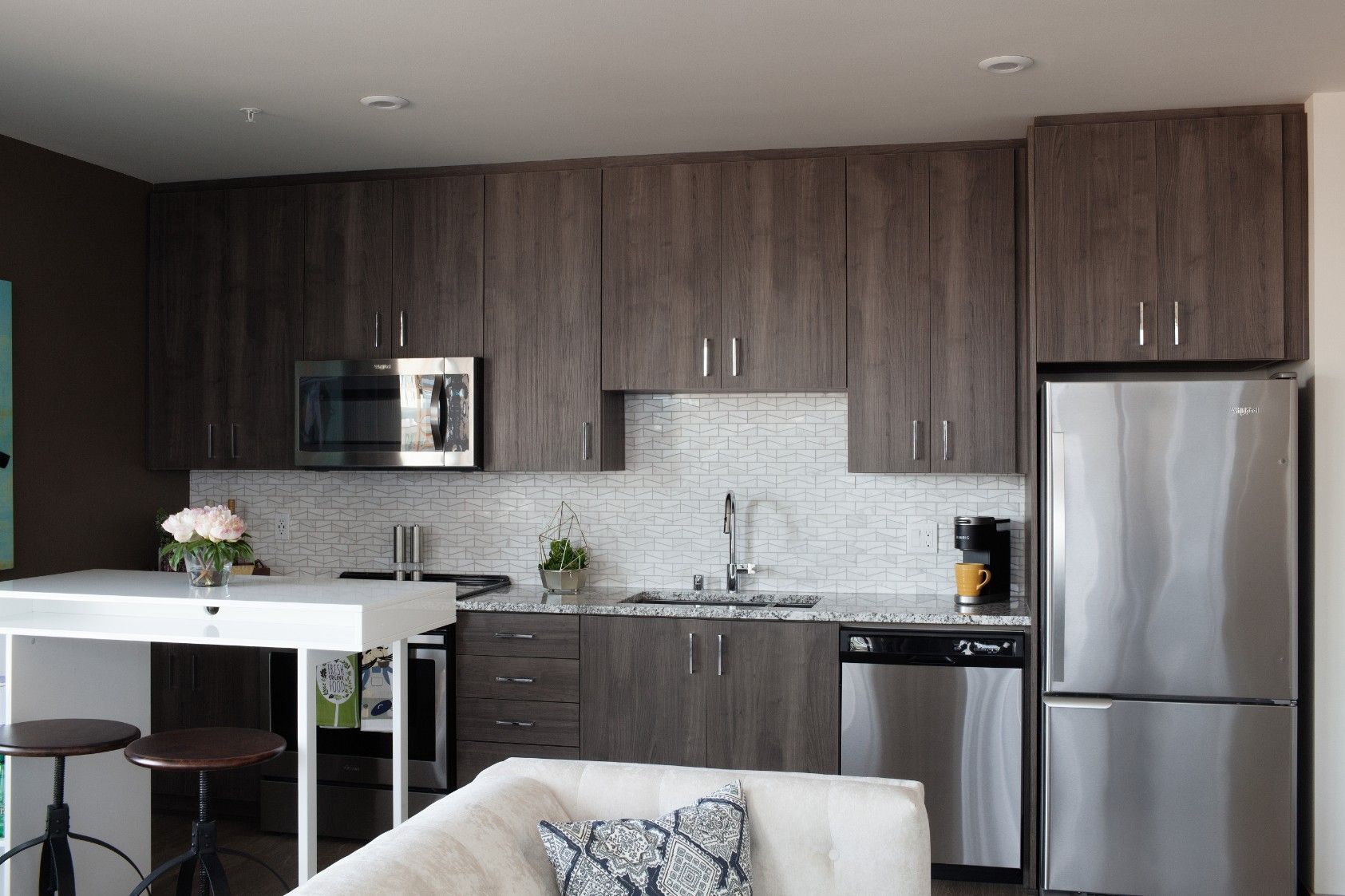 Modern kitchen and living area with dark wood cabinets, stainless appliances, white island, and a sofa in the foreground