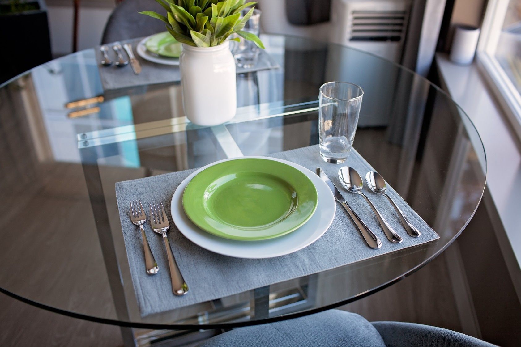 Set dining table with green plates, silverware, glassware, and a small potted plant on a round glass table.