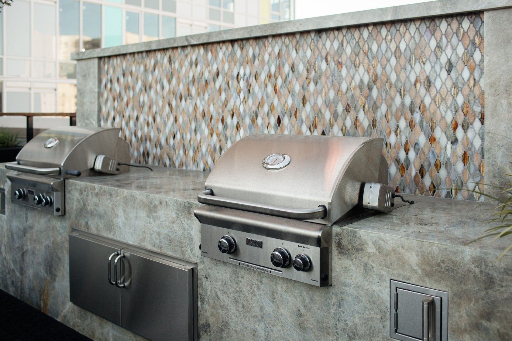 Two stainless steel grills built into a concrete outdoor counter against a patterned tile wall