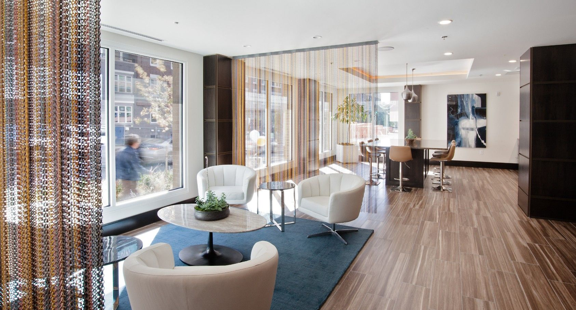Bright modern living room with blue rug, white chairs, sheer curtains, and city view through large windows
