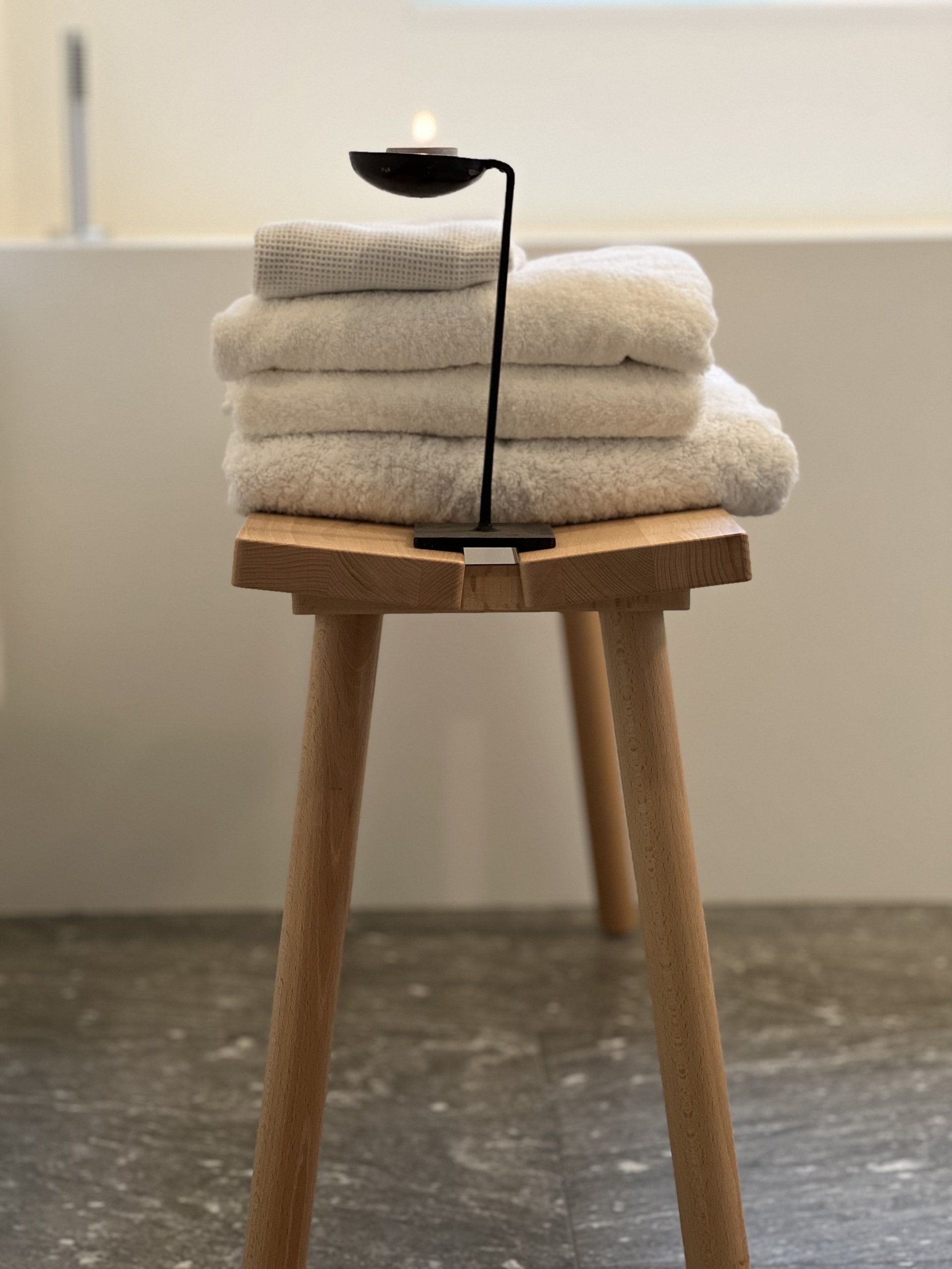 Een stapel witte handdoeken en een kaars op een klein houten krukje in een badkamer.