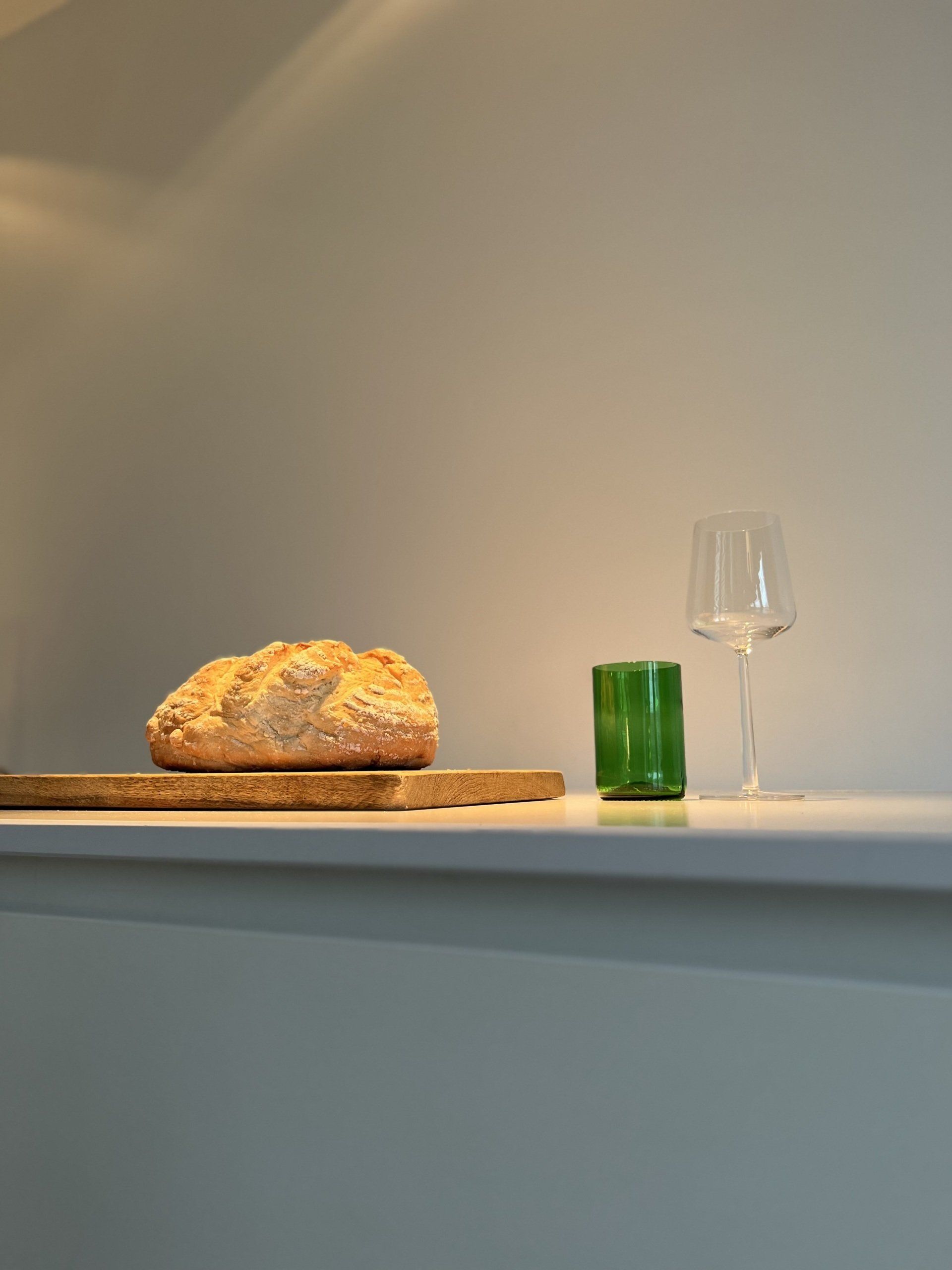 Een brood op een snijplank, een groen glas en een wijnglas op een wit aanrechtblad tegen een neutrale muur.