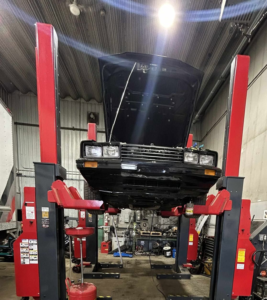 Voiture noire soulevée sur un pont élévateur hydraulique rouge et gris dans un garage, capot ouvert.