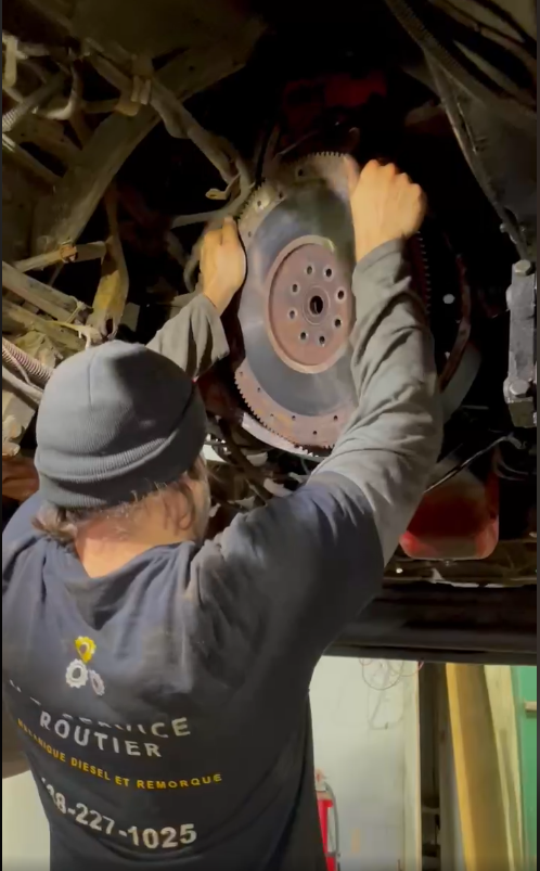 Un mécanicien installe un gros volant moteur en métal sous un véhicule ; l'espace de travail est faiblement éclairé.