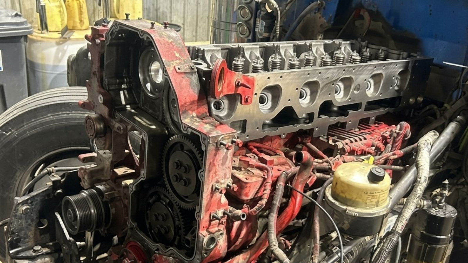 Compartiment moteur endommagé d'un camion rouge, avec tuyaux et câbles apparents, vu de côté pendant les réparations.