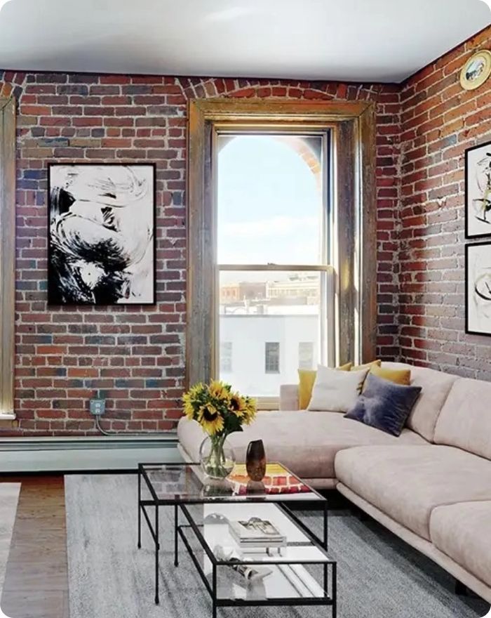 Loft living room with exposed brick walls, beige sectional sofa, glass coffee table, and large window