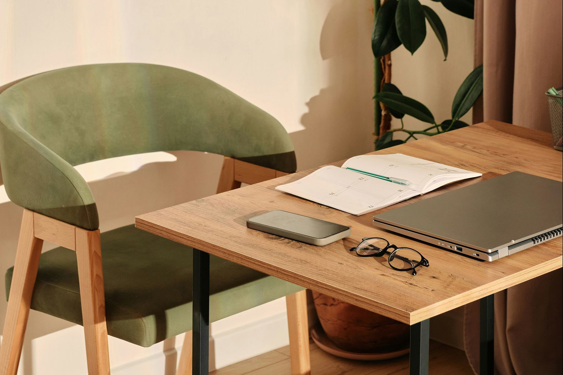A wooden desk with a green chair, laptop, notebook, phone, keys, and potted plant by a window.