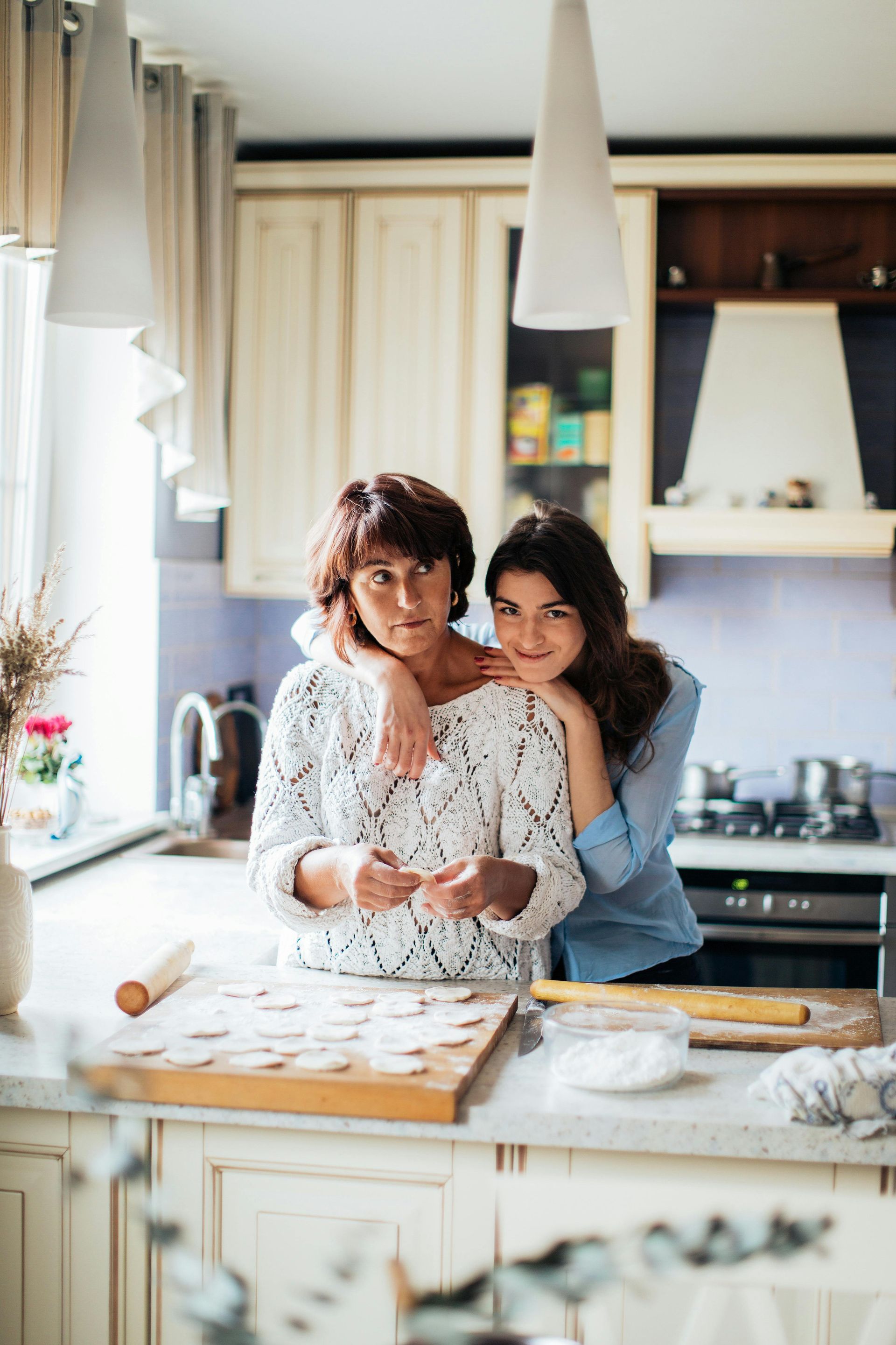 Two people stand in a kitchen, one hugging the other from behind while preparing dough on a wooden cutting board.