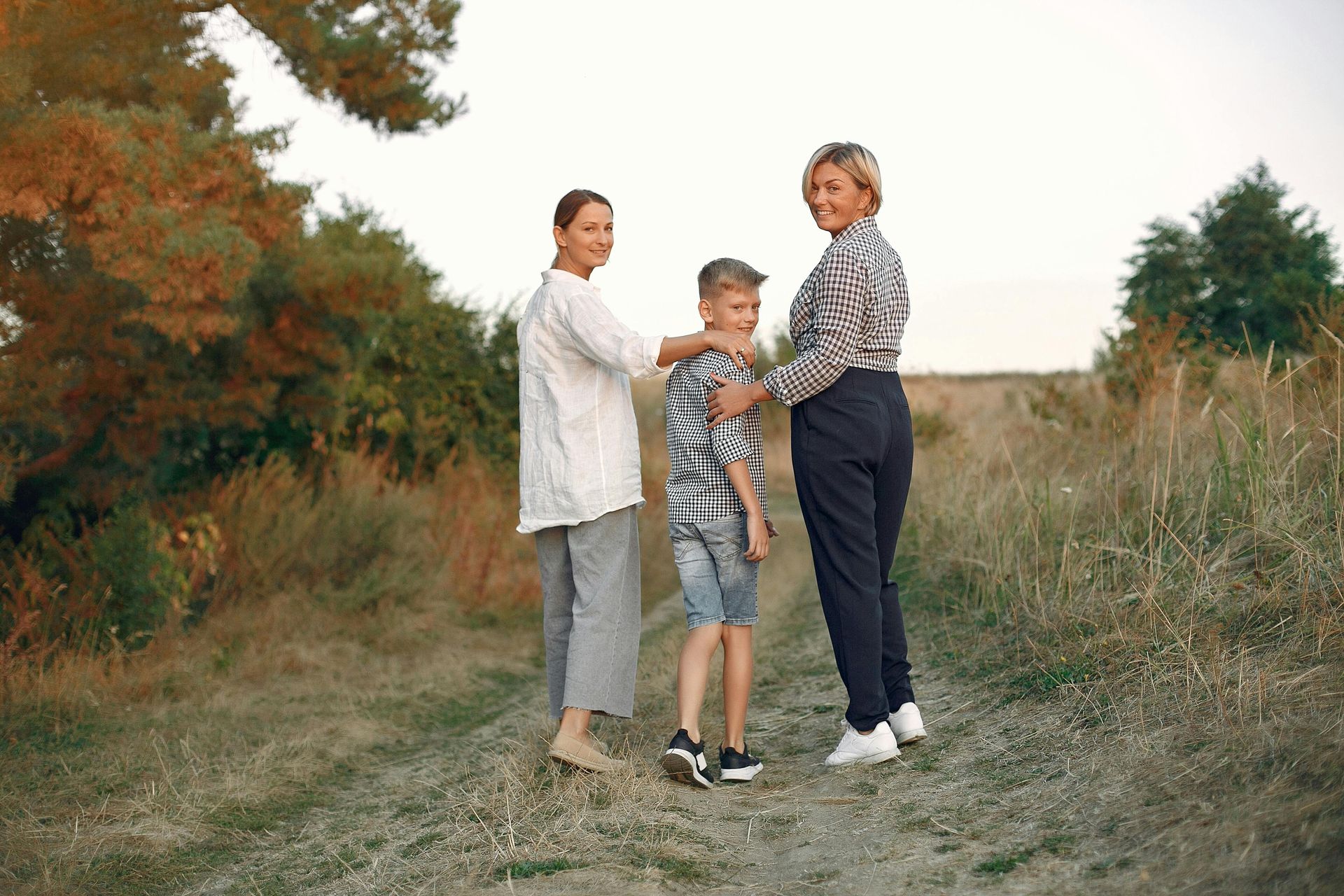 Two adults and a child walking on a dirt path in a rural landscape, looking back at the camera.