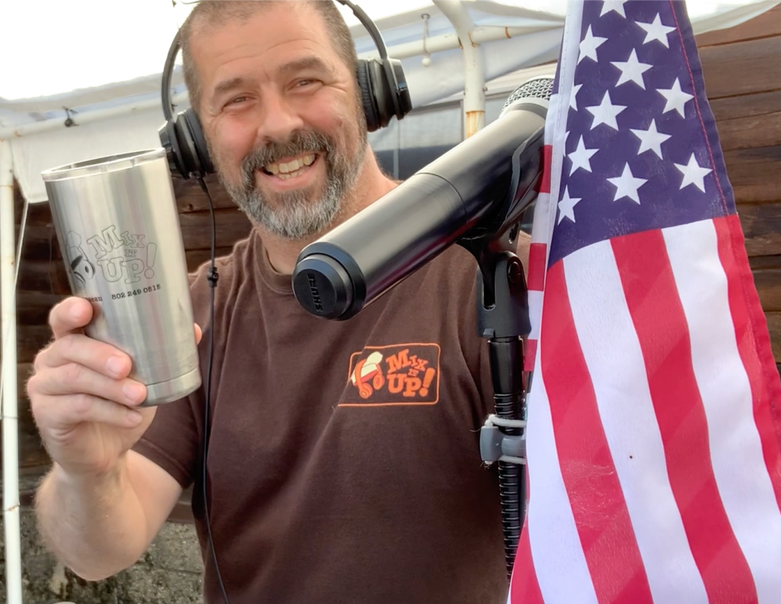 DJ holding up a Mix It Up! coffee mug, while wearing a head set. With his microphone and American Flag beside him