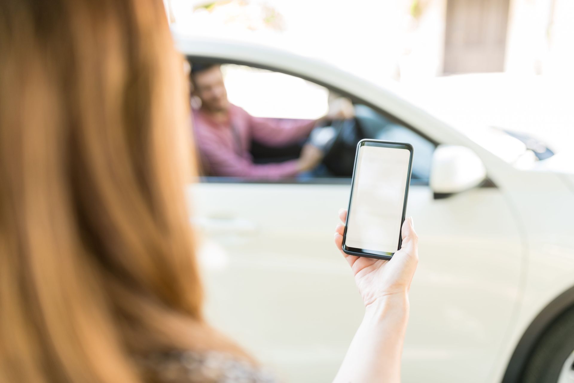 Woman holding a smartphone near a white car with a driver visible through the window