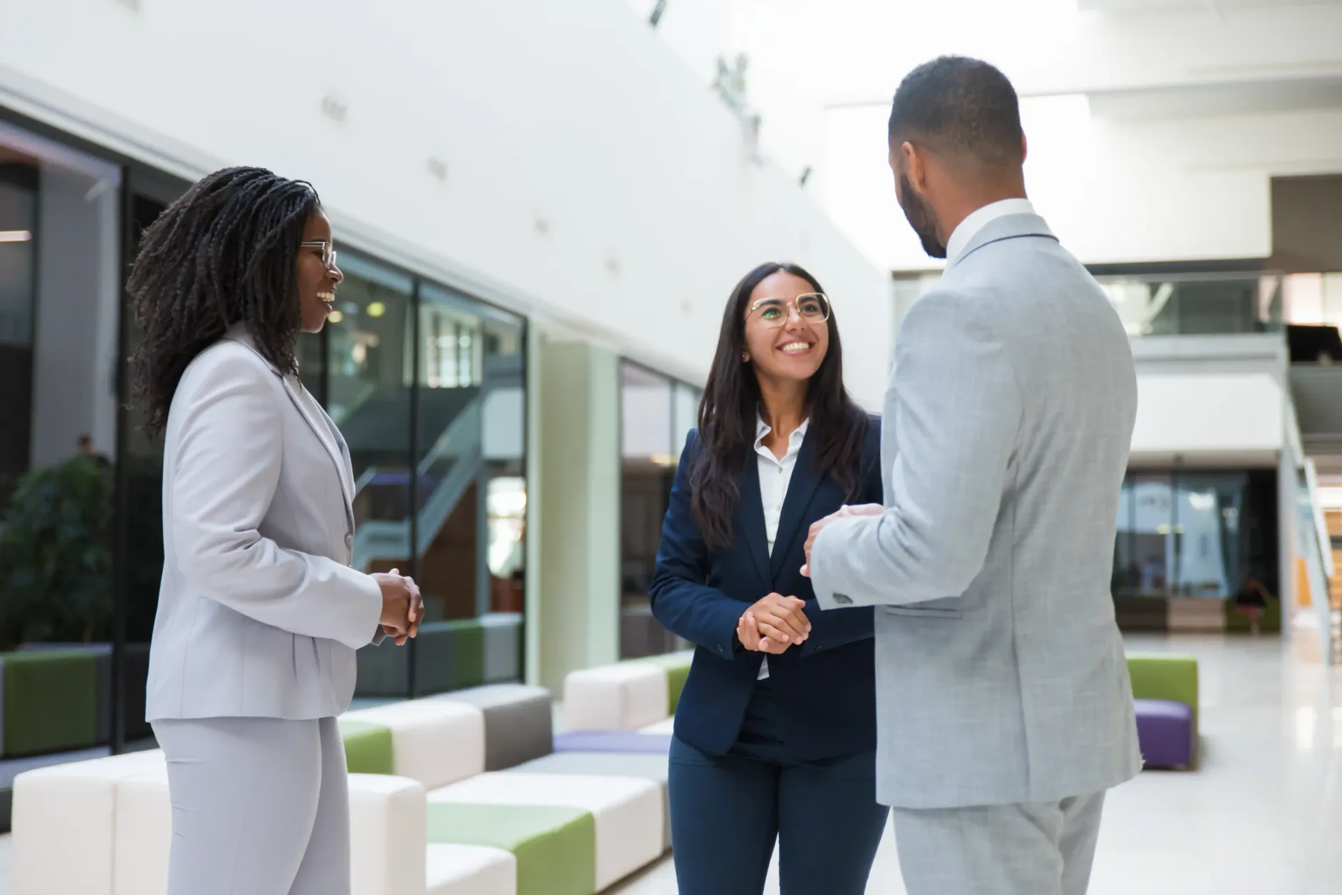 Three coworkers talking in a bright office lobby, one woman smiling in a navy suit.