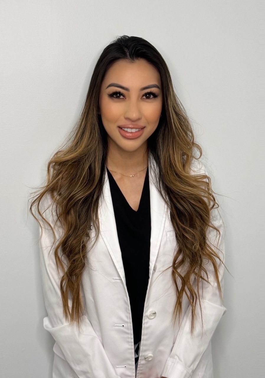 Young woman in a white lab coat smiles, long brown hair, against a light gray backdrop.