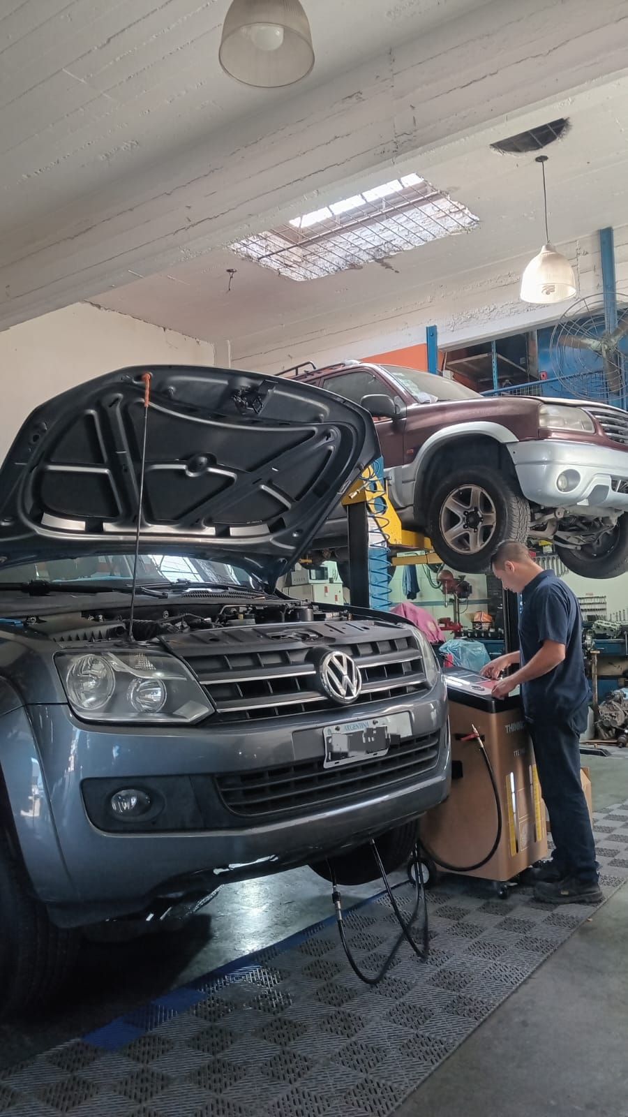 Coche en un garaje con el capó abierto, mecánico trabajando junto a piezas y equipos apilados.