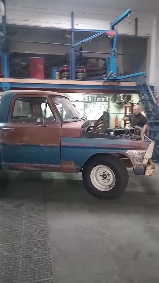 Camioneta vieja azul y oxidada en un garaje, con el capó abierto y una persona trabajando dentro.