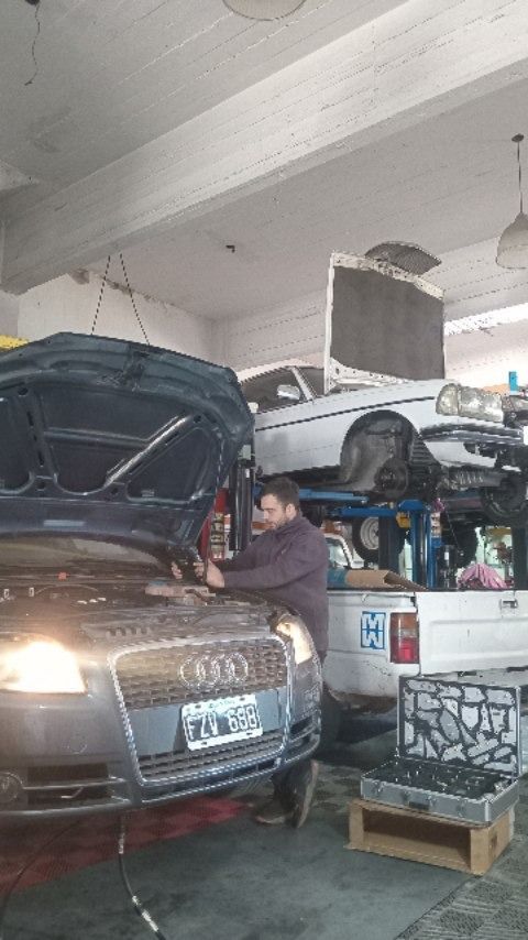Coche en un garaje con el capó abierto, mecánico trabajando junto a piezas y equipos apilados.