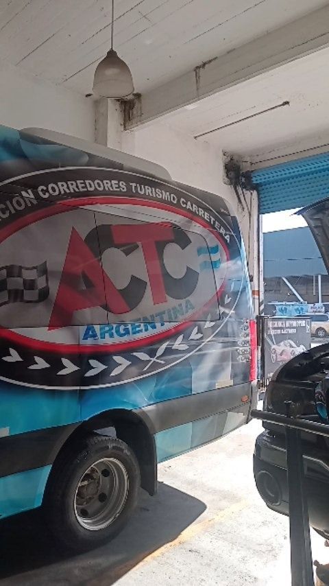 Un camión con remolque de la empresa ACT Argentina, de color rojo y negro, estacionado bajo una acera techada cerca de un edificio.