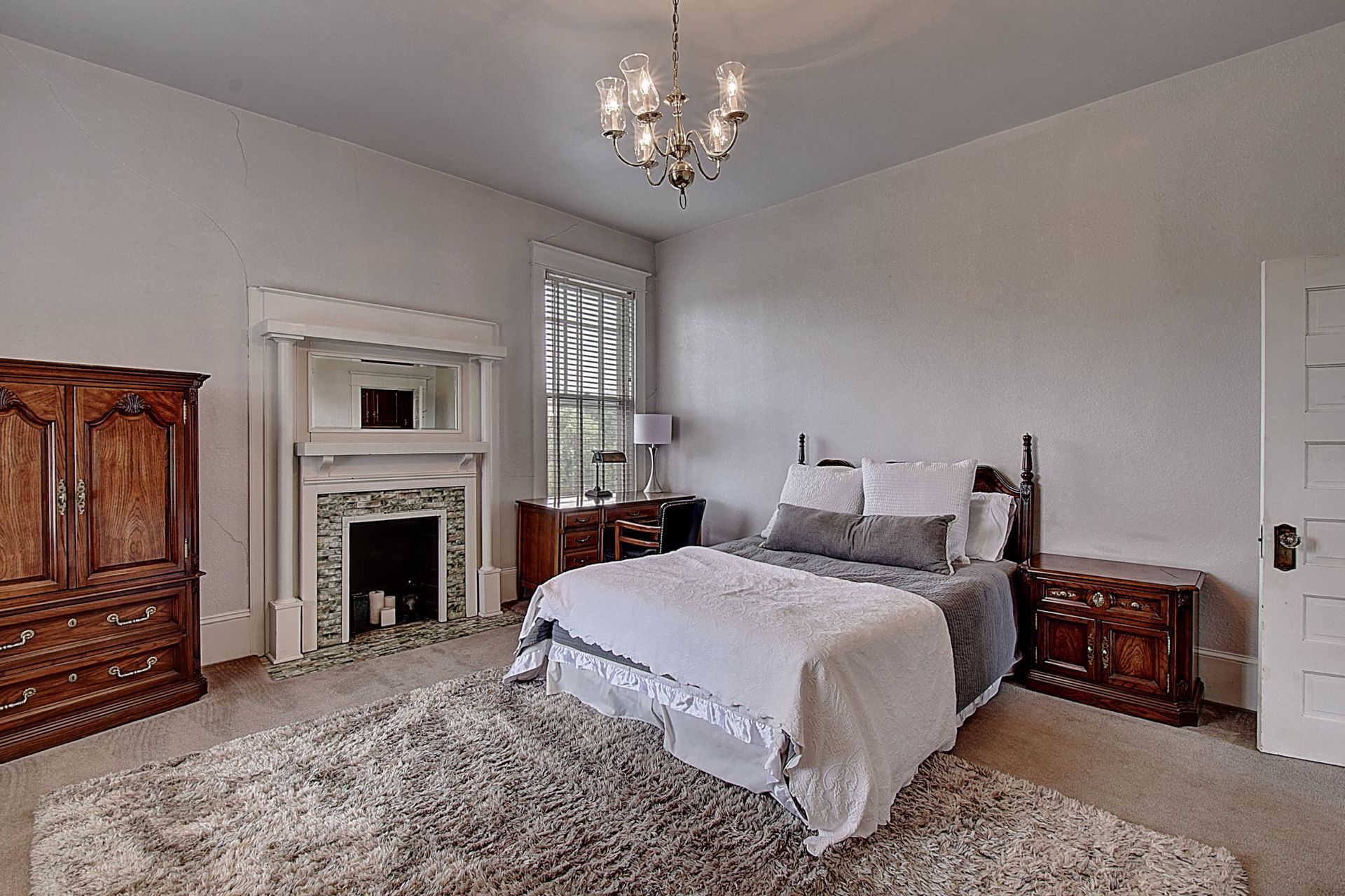 A bedroom with a bed , dresser , nightstand , fireplace and chandelier.
