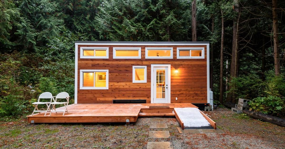 A small wood cabin with multiple windows. There are two lawn chairs set up on the porch and the cabin is surrounded by trees.