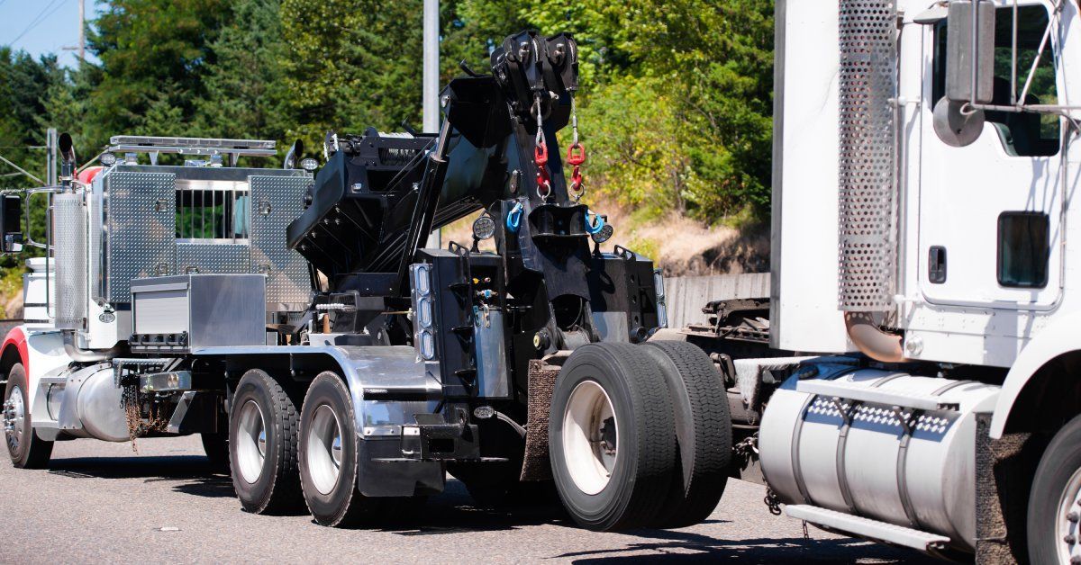 A large red and silver big rig towing another white semi truck tractor on a road. The road is surrounded by trees.