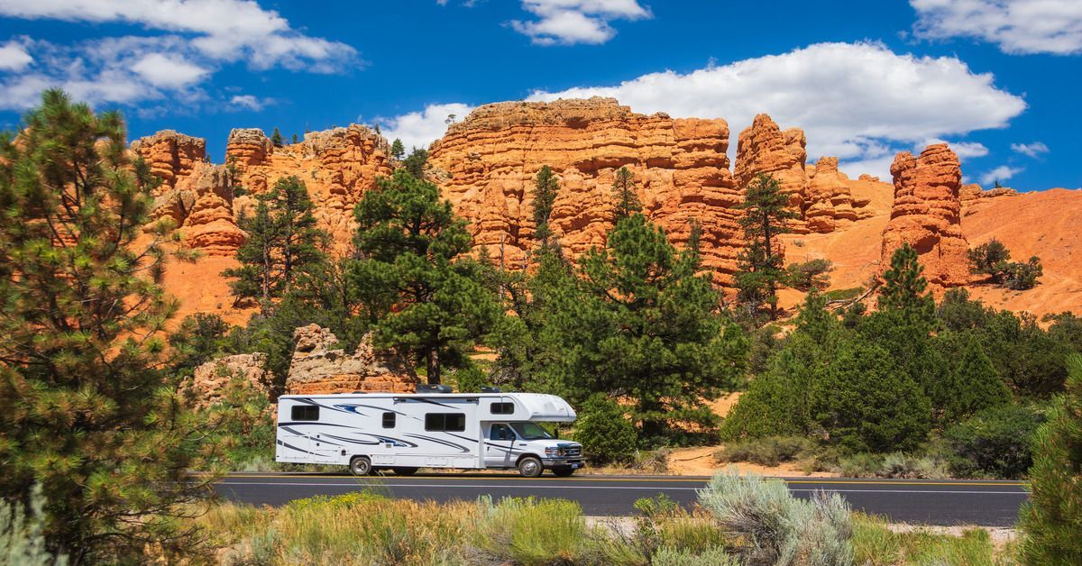 A large, white colored RV driving on a road surrounded by a background of tall, red canyons and multiple pine trees.