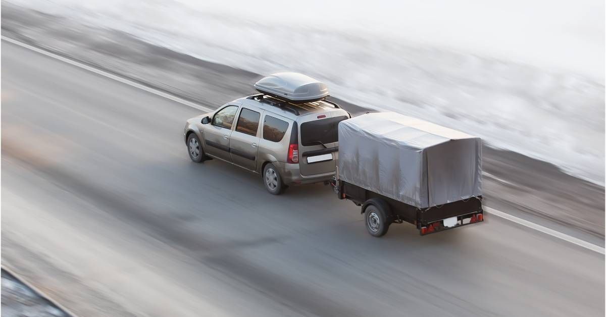 A gray SUV pulling a trailer and driving along a highway road surrounded by snow. The road around the vehicle is blurry.