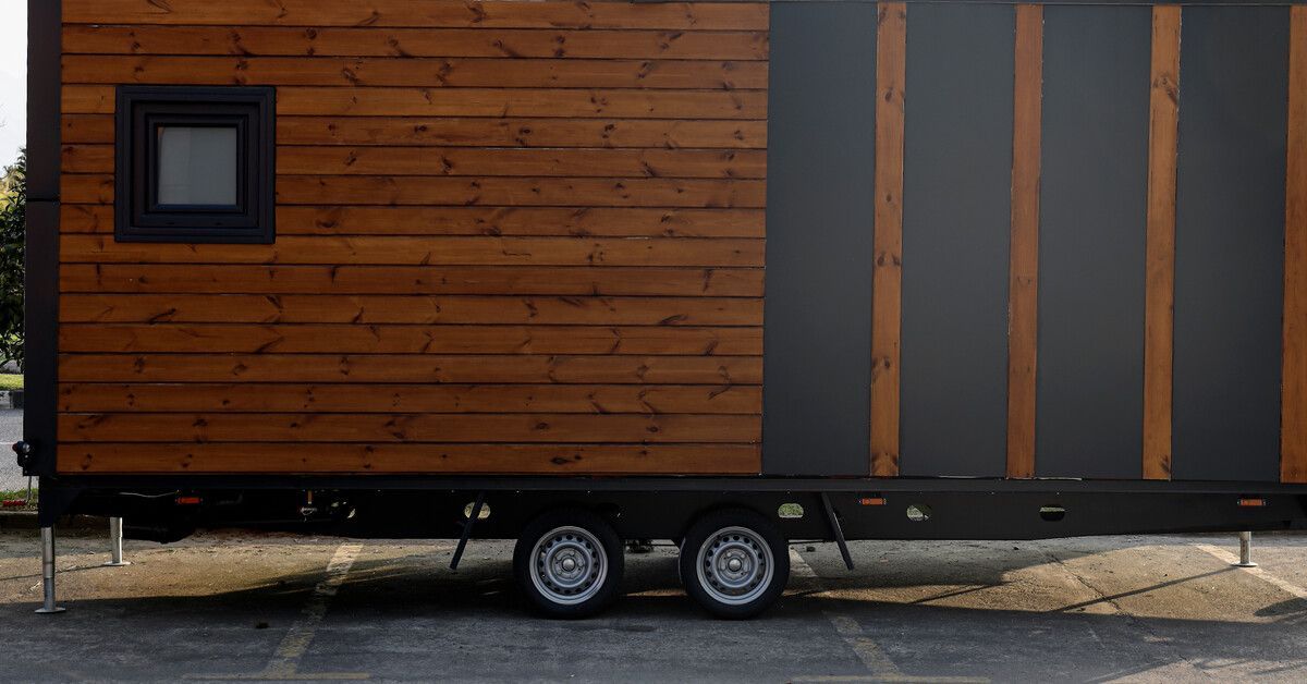 A tiny mobile home made out of wood is parked in a parking lot. Attached to the home are wheels and a trailer hitch.
