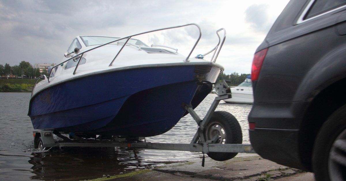 A black car uses a trailer hitch to pull a blue and white motorboat out of a lake and onto the pavement.