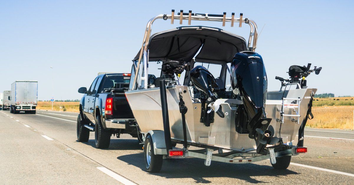A black pickup truck tows a silver boat down an interstate highway behind a couple of semitrucks on a sunny day.