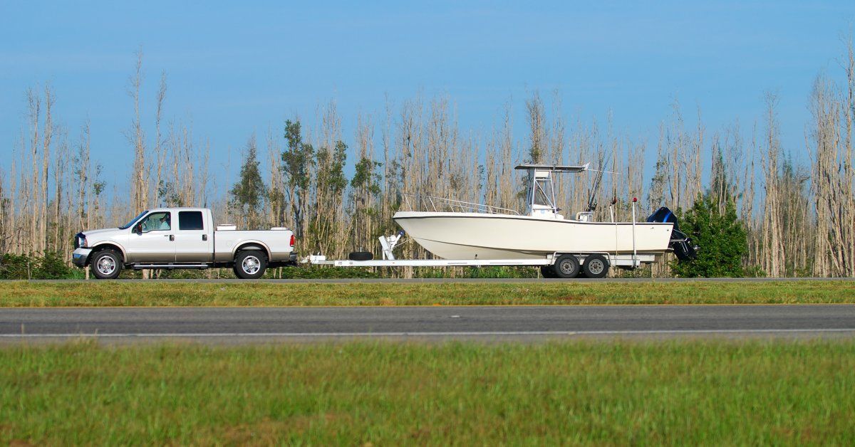 A white pickup truck drives down a highway while using a trailer hitch to tow a white boat behind it.