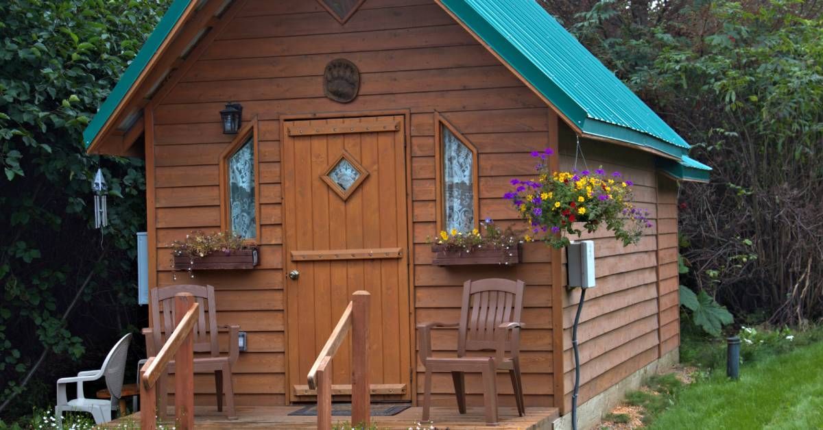 A small, brown, wooden house with a green roof near a background of trees. Plants, flowers, and chairs decorate the porch.