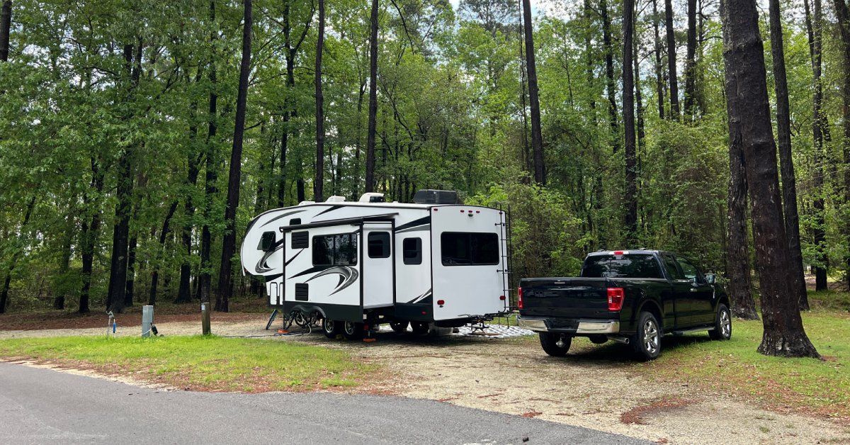 A white and black RV is parked on a dirt plot in the middle of a forest. A black pickup truck is parked next to it.